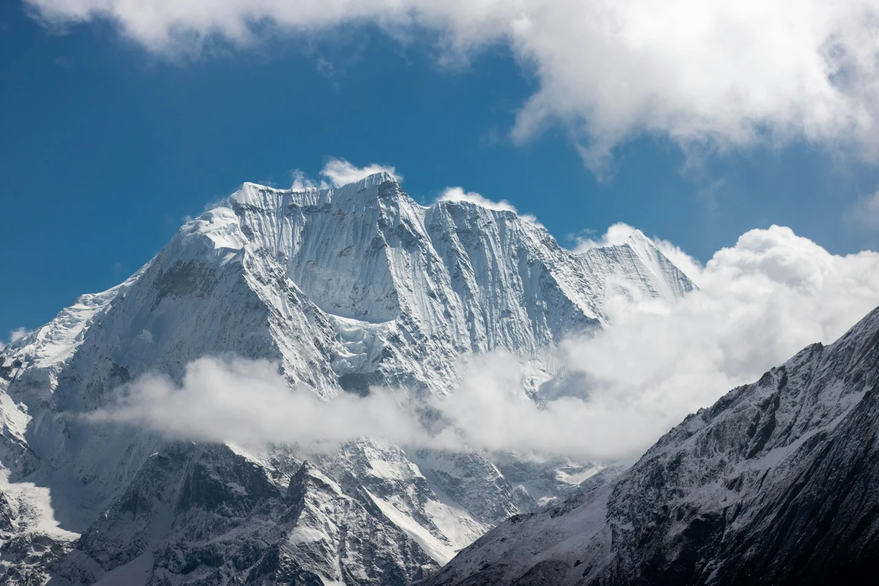 A panoramic view of the snow-covered Mount Manaslu peak in the Himalayas of Nepal, partially shrouded by white clouds under a clear blue sky. Another rugged, dark mountain ridge is visible in the lower right foreground.