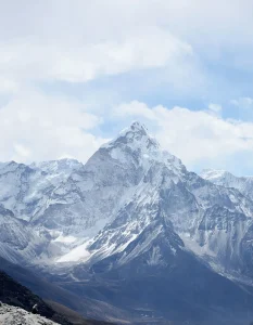 A snow-covered, jagged mountain peak rises sharply toward a cloudy blue sky. Steep, rocky ridges and smaller snowy slopes surround the prominent central summit.