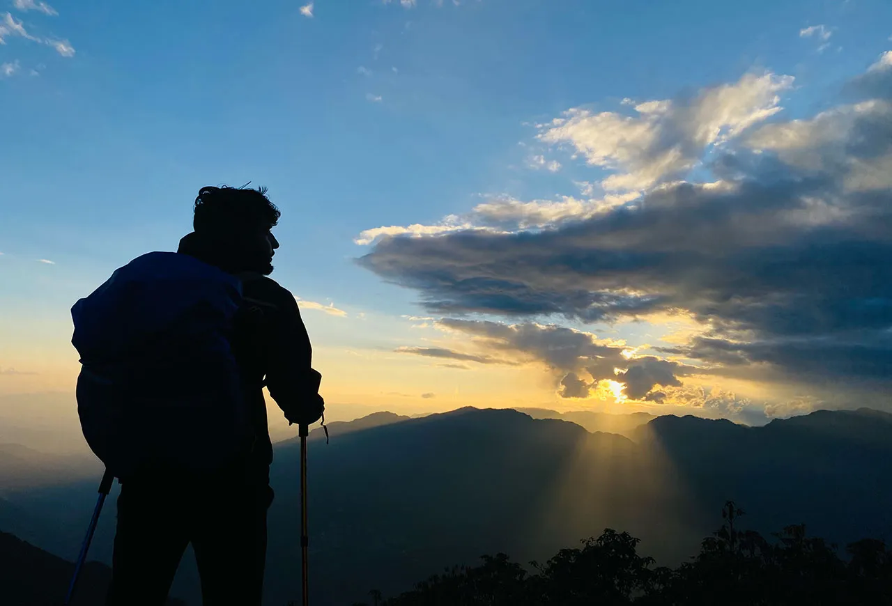 Silhouette of a hiker with a backpack and trekking poles overlooking a dramatic mountain sunset with sunbeams through clouds.