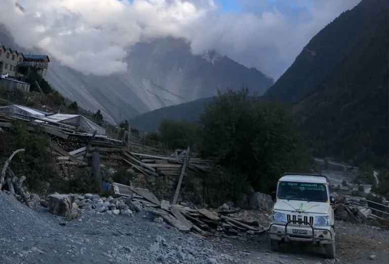 White SUV parked on a rugged mountain road in a Himalayan village with cloudy peaks in the background.