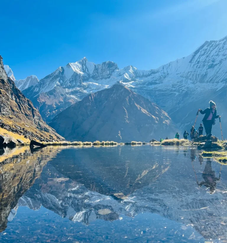 A group of trekkers with hiking poles reflecting in a crystal-clear mountain lake, with the snow-covered peaks of the Annapurna Massif in the background under a bright blue sky.